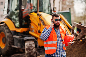 Obraz premium Brutal beard worker man suit construction worker in safety orange helmet, sunglasses against traktor with mobile phone at hand.
