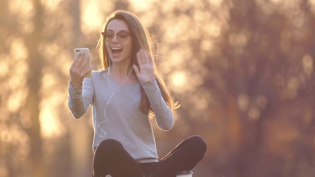 Happy young woman making videocall with phone and headphones and waving hand in park