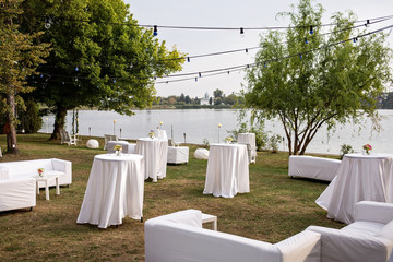 White tables outside by the lake for a wedding party, Green trees by the lake with white tables and lounge couches, Outside party by the lake