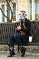 Fototapeta na wymiar Emotional portrait of happy beautiful woman with red braids hair and natural makeup wearing black jeans trench grey sweater surfing on her smartphone in spring city park.