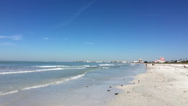 Small waves roll ashore along St Pete Beach, Florida as beachcombers stroll in the distance looking for seashells.