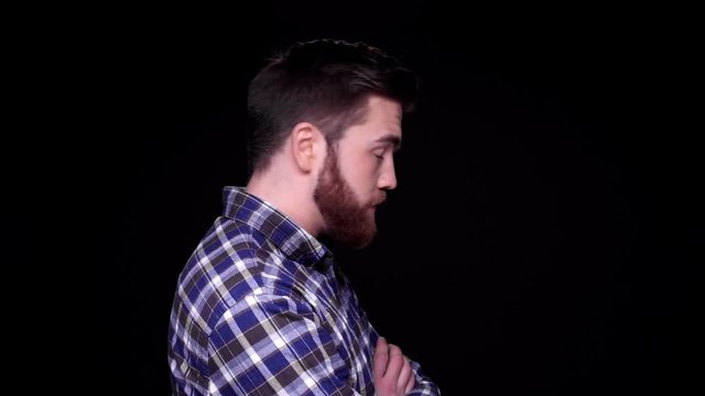 Grumpy offended young bearded man standing with arms folded and looking at camera isolated over black