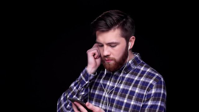Young casual bearded man in earphones choosing song on mobile phone isolated over black background