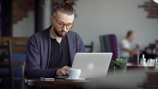 A young attractive hipster in a cafe, with a modern interior. The man works on his stylish laptop. A designer or a writer makes a project in a cafe.