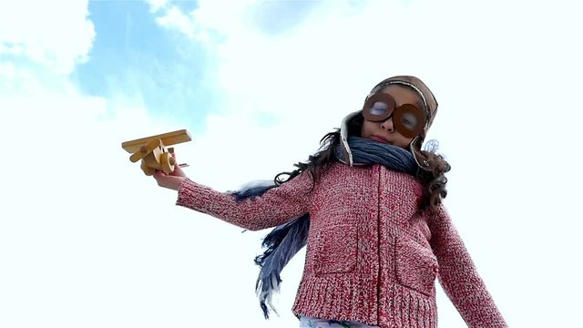 Little girl with pilot hat playing with wooden airplane against blue sky, slow motion