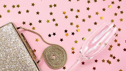 Festive evening golden clutch and champagne glass with star sprinkles shining on pink. Holiday and celebration background. Luxury accessories and party concept.