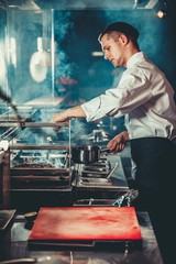 Food concept. Chef in white uniform monitors the degree of roasting and greases meat with oil in saucepan in interior of modern restaurant kitchen. Preparing traditional beef steak on barbecue oven.