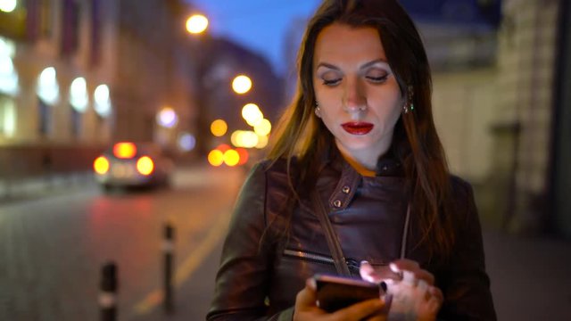 Woman with red lips uses smartphone and strolls along the medieval street at everning