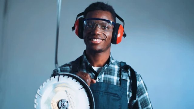 Young African-American man construction worker with angle grinder machine wearing safety ear muffs and checked shirt with overalls uniform, standing against white wall and smiling
