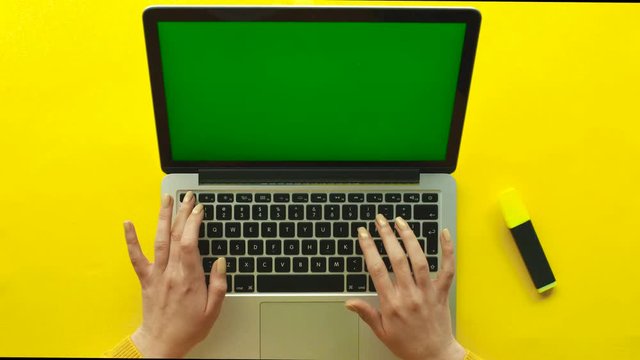 Woman hands typing on laptop keyboard on yellow table background. Laptop with green screen. Top view. Chroma Key