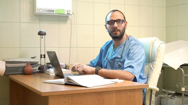 Happy male doctor with laptop talking good news sitting by table in office
