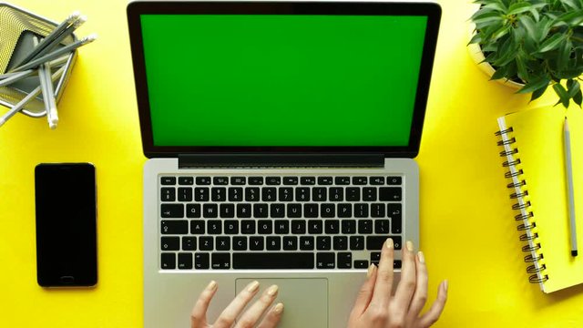 Woman hands typing on laptop keyboard on yellow table background. Laptop with green screen. Top view. Chroma Key