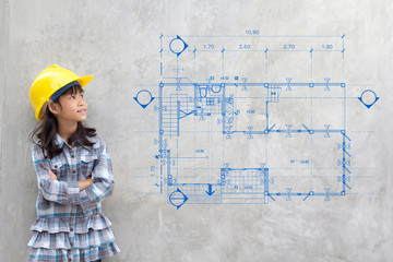 building, childhood, developing, construction and architecture concept - smiling little girl in yellow helmet showing blueprint drawing on wall