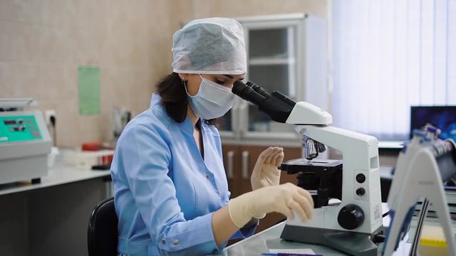 Professional female laboratory assistant in blue lab coat, protective mask, cap and gloves sitting in cabinet and examining medical samples under the microscope. Woman in uniform is working with