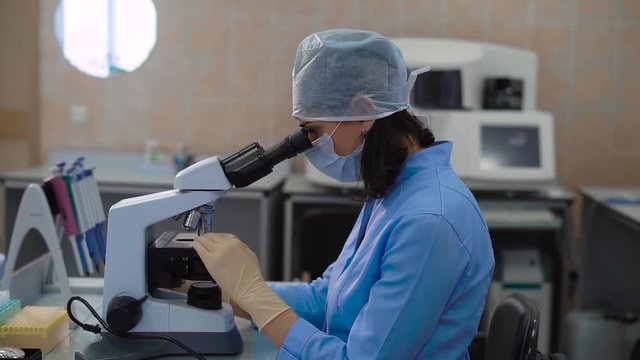 Female medical research assistant in blue lab coat working in laboratory and examining samples under the microscope. Woman in uniform analyzing specimen with high concentration.