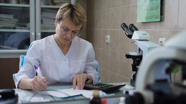 Professional female research assistant in white coat sitting at the table with microscope and calculator in front of her processing medical data. Doctor in uniform working with results of examination.