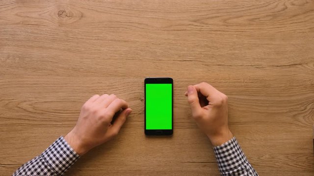 Man using smart phone with green screen on wooden table background. Male hands scrolling pages, zooming, tapping on touch screen. top view. Office desk background. Chroma key.