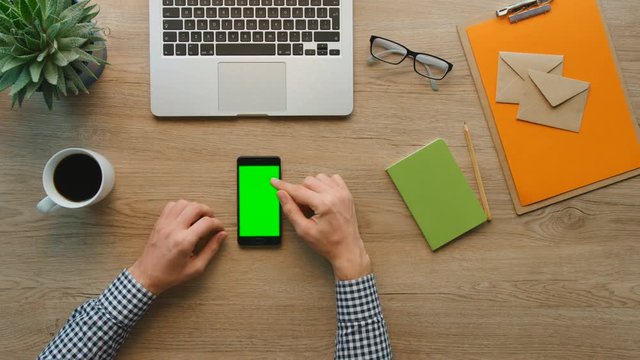 Man using smart phone with green screen on wooden table background. Male hands scrolling pages, zooming, tapping on touch screen. top view. Office stuff on desk background. Chroma key.