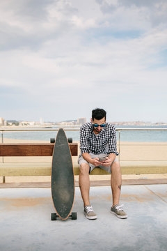 Young handsome bearded man in sunglasses sitting on bench near skateboard and using his smartphone.
