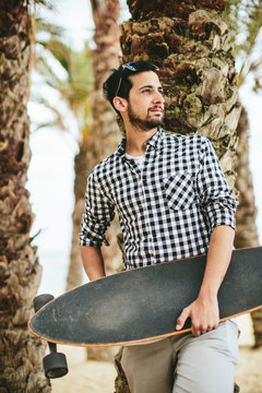 Young smiling man with skateboard leaning on palm tree.