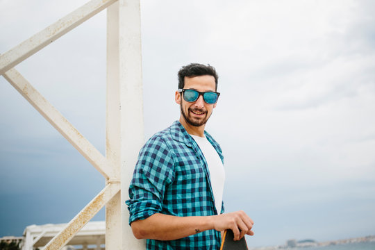 Handsome Bearded Man In Summer Outfit Standing On The Shore With Skateboard And Smiling.