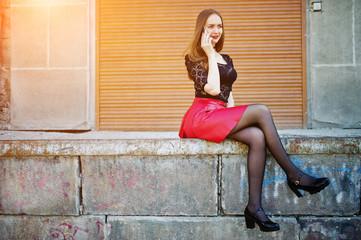 Portrait of girl with black choker on her neck, red leather skirt and mobile phone at hand against orange shutter.