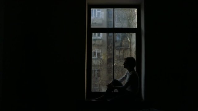 Silhouette of depressed woman sitting on the sill and looking out the window