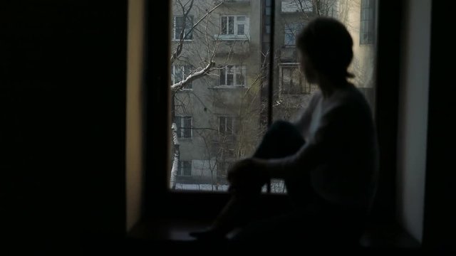 Silhouette of depressed woman sitting on the sill and looking out the window