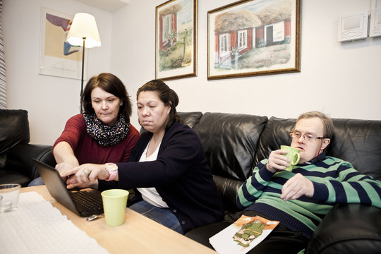 Healthcare worker assisting woman with down syndrome in using laptop while man having drink at sofa