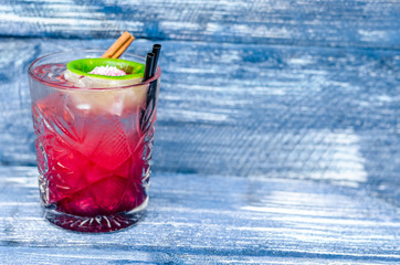 A glass of red cocktail with ice, lime and straw, on a blue denim background. Side view