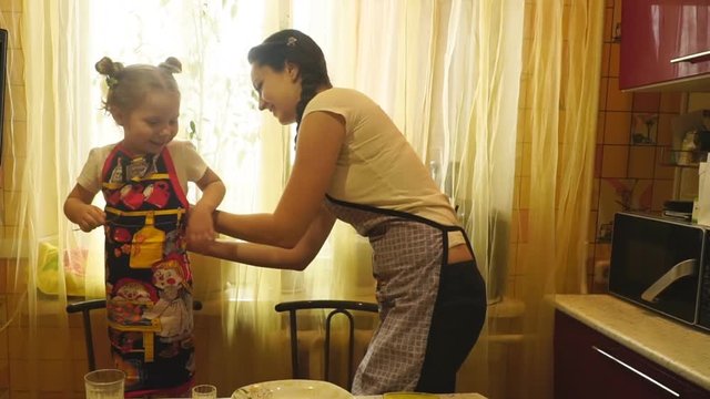 Mother tying an apron adjusts her daughter in the kitchen