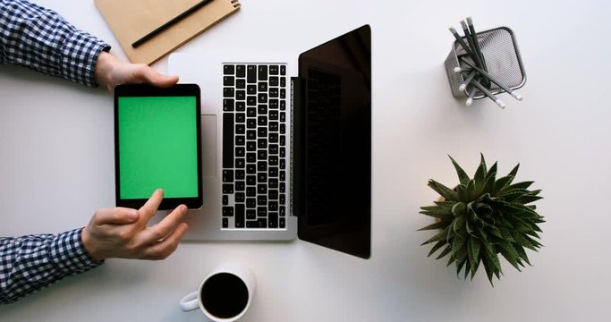 Top view of man working on laptop and black tablet device with green screen on wite office table. Scrolling pages, tapping on touch screen. Chroma key