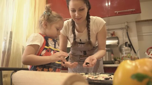 The girl and her mother in the kitchen is applied on the raw pizza toppings