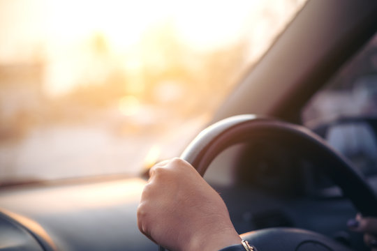 Woman's Hands Holding On Black Steering Wheel While Driving A Car During Sunset