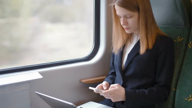 Young Beautiful Business Lady is Traveling by the Window in the Train and Working on her Laptop and Texting Message at the Mobile Phone