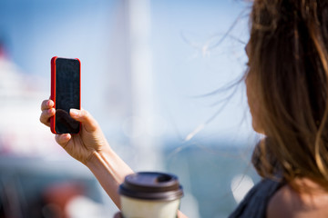 Beautiful woman taking  photo with smart phone. Picture of harbor with sailboats, seaside. Sunny...