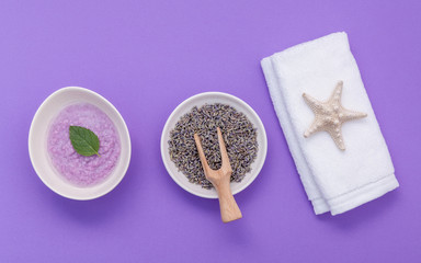 Homemade skin care lavender bath salt beauty treatment, towel and lavender flower on purple background flat lay.