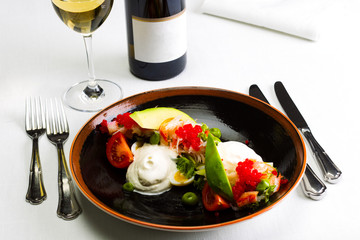 Seafood salad with red caviar and vegetables with a bottle and a glass of wine on a white tablecloth background, studio light
