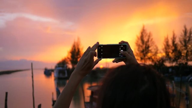 Young Mixed Race Girl Taking Photo of Beautiful Sunset Using Mobile Phone at Fisherman Pier. HD Slowmotion. Thailand.