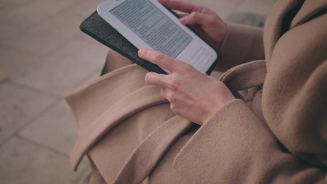 Young woman in pastel beige colours warm autumn coat reads a white ebook on a cold and fresh morning in a soft light, turns the pages in a calm and relaxed manner on a loop