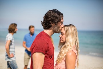 Fototapeta na wymiar Man kissing girlfriend on forehead against friends at beach