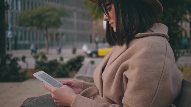 A beautiful brunette student in trendy glasses rests in tranquility while reads through the imaginative and overwhelming novel on her ereader while the world moves around her on a fast pace.