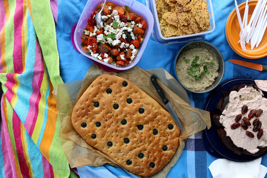 Top view of various picnic food: roasted vegetable and feta salad, baba ghanoush, gluten-free crackers, foccacia, gluten-free and sugarfree dates cake.