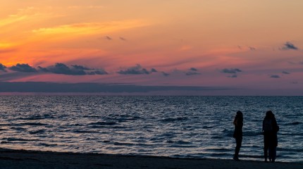 Naklejka na ściany i meble Jurmala (Riga), Latvia - April 16, 2017: Golden sunset on the Baltic sea with silhouettes of people (shadows). Coastal landscape with setting Sun reflections on the water and sand (in Latvia)