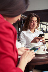 Smiling and serious gorgeous women surfing on Internet in smartphones during lunch with cup of coffee