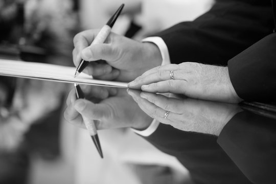 foto b/n di una mano che firma con una penna un  documento - riflette sul tavolo