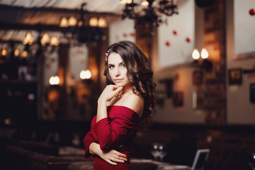 Plakat Beautiful European girl in a long cocktail evening dress of red coral with curly hair is standing in the restaurant. Concept of the make-up artist's work.