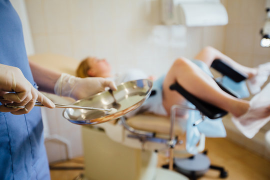 Red hair female doctor holding tool in steel basin in front of the female patient on gynecological chair. Close up