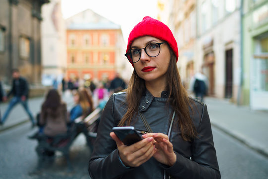 Woman with red lips and red hat uses smartphone and strolls along the medieval street