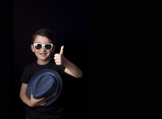 Low key portrait of young boy isolated on black background. Plenty of space left for copy. Holding hat, wearing sunglasses